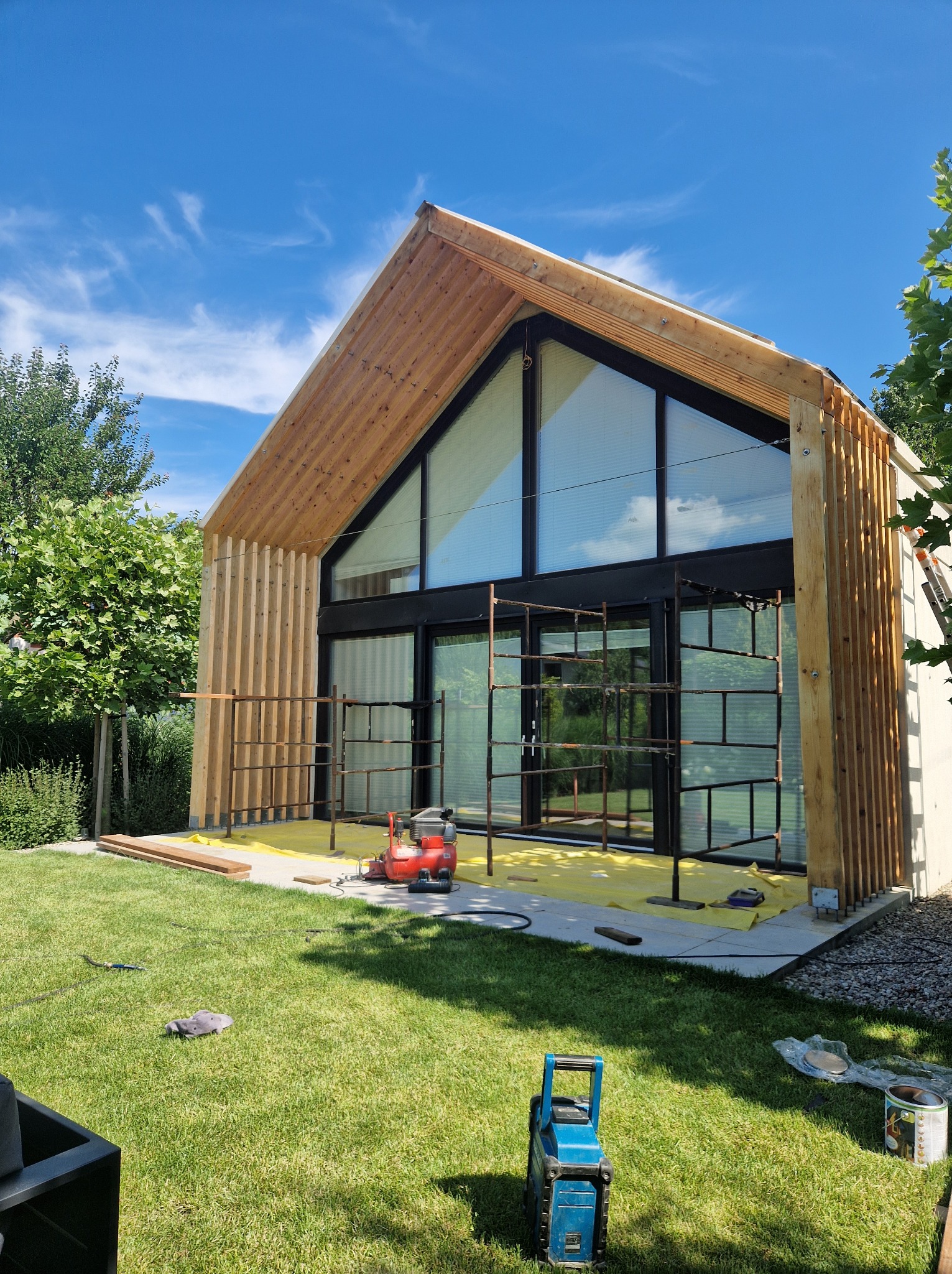 Wznoszenie konstrukcji z drewna na zewnątrz budynku z dużymi oknami, widoczne rusztowanie, kompresor i narzędzia na trawniku