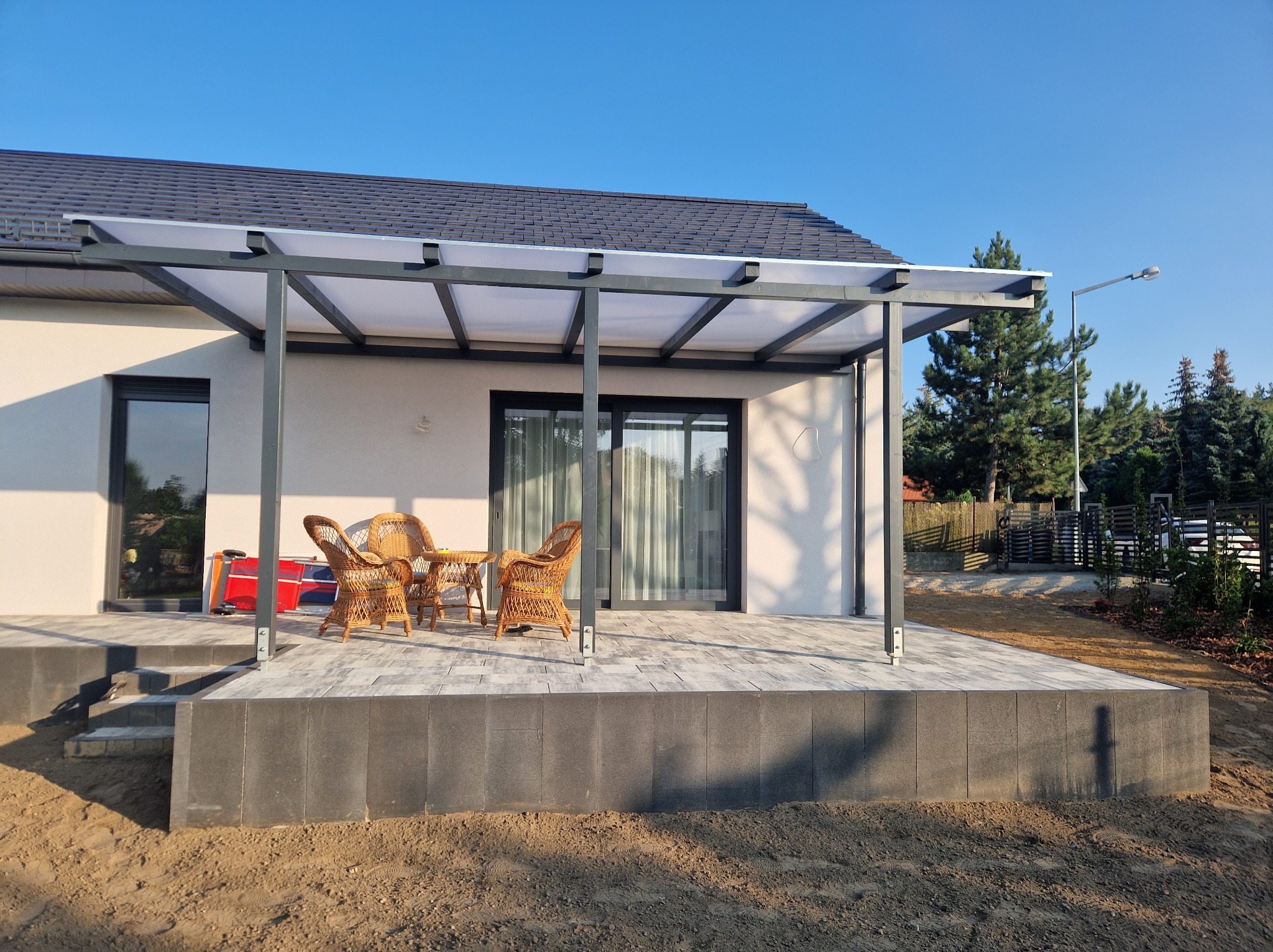 Nowoczesny taras z szarych płyt, zadaszony konstrukcją z poliwęglanu, wyposażony w wiklinowe krzesła i stolik, widok na ogród i błękitne niebo.