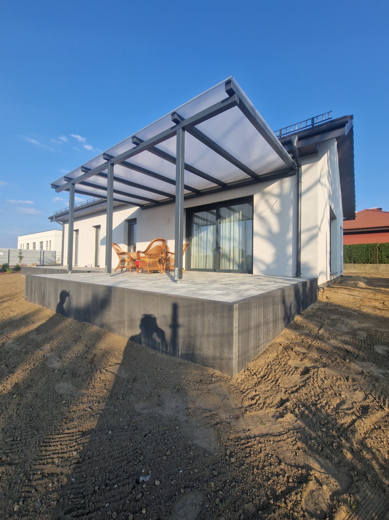 Nowoczesny dom z zadaszonym tarasem wyłożonym szarą kostką, wyposażonym w wiklinowe fotele, na tle błękitnego nieba i przygotowanego terenu pod ogród.