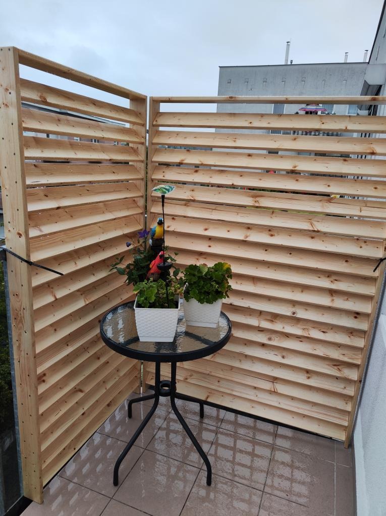 Balkon z drewnianą przesłoną lamelową, na której tle stoi mały, okrągły stolik z dwoma doniczkami z kwiatami.