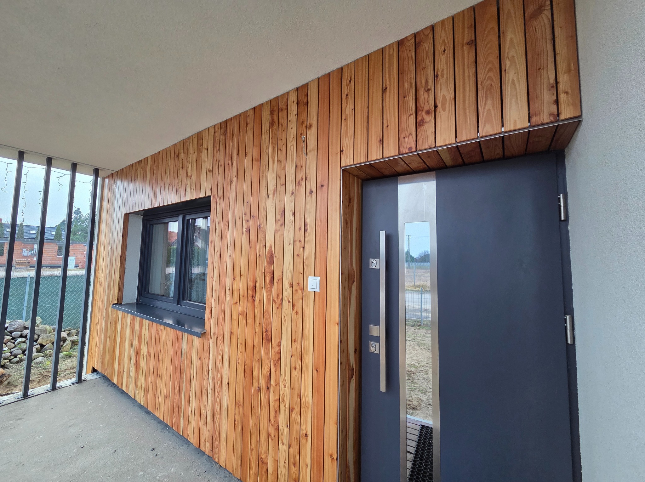 Elewacja budynku z modrzewiową okładziną pionową, szare okno i drzwi wejściowe z długim, pionowym uchwytem.