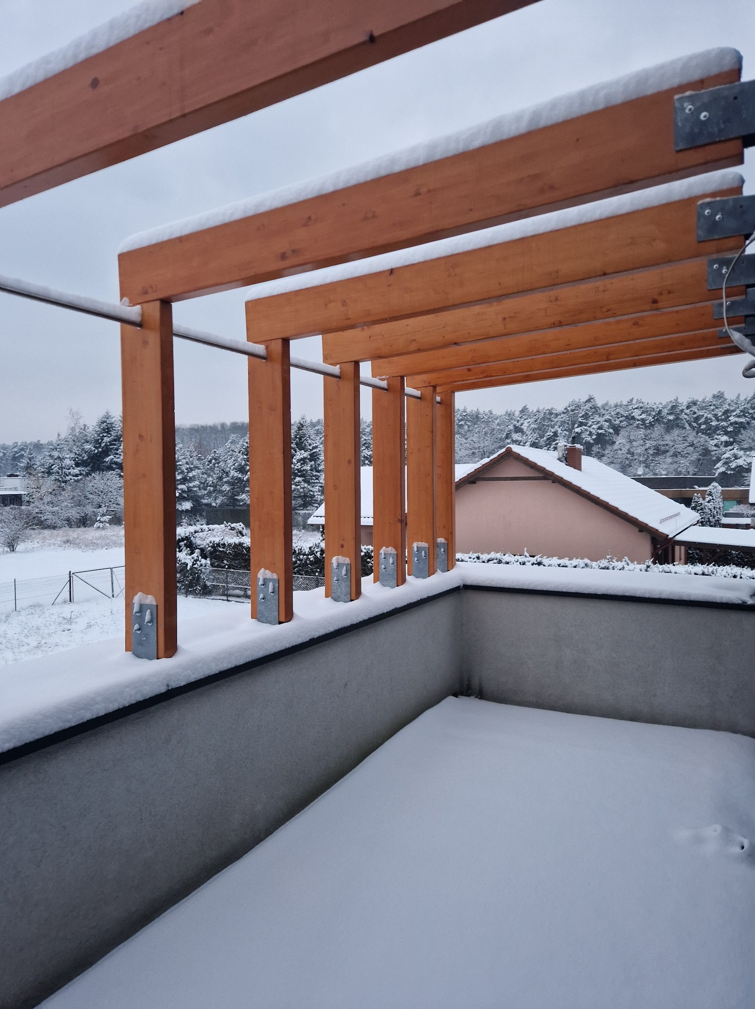 Zimowy widok z balkonu z drewnianą konstrukcją zadaszenia, pokrytą śniegiem, na tle zasypanego śniegiem krajobrazu z domami i lasem.