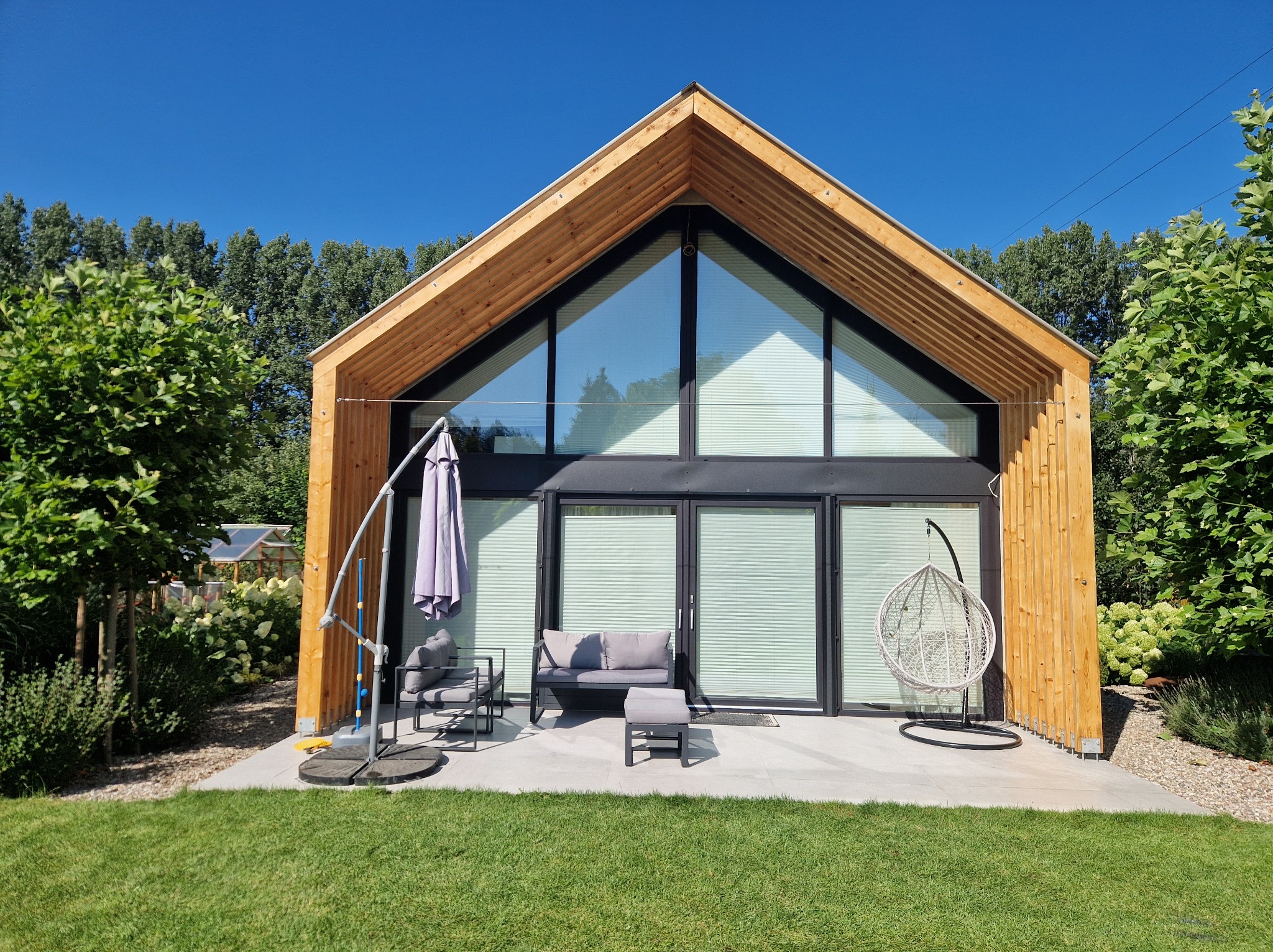 Nowoczesny dom z drewnianą elewacją i dużym przeszkleniem, taras z szarymi meblami ogrodowymi i wiszącym fotelem, zadbany trawnik, błękitne niebo.