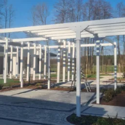 Nowoczesna, biała pergola metalowa z pionowymi elementami, na tle domu i drzew. Kostka brukowa na podłożu. Słoneczny dzień, błękitne niebo.