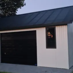 Nowoczesny garaż blaszany z czarną bramą segmentową i dwuspadowym dachem, oświetlony wieczornym światłem, na tle zieleni. Widoczne okno z brązową ramą.