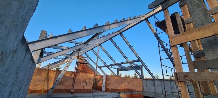 Konstrukcja stalowa dachu w trakcie budowy, widok z dołu na metalowe elementy i ceglane ściany, niebo w tle. Widoczne rusztowania drewniane i drabina.