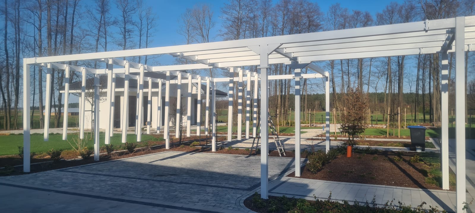 Nowoczesna, biała pergola metalowa z pionowymi elementami, na tle domu i drzew. Kostka brukowa na podłożu. Słoneczny dzień, błękitne niebo.