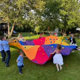 Grupa dzieci bawi się kolorową chustą animacyjną, podrzucając w górę kolorowe piłeczki na trawniku podczas imprezy plenerowej.