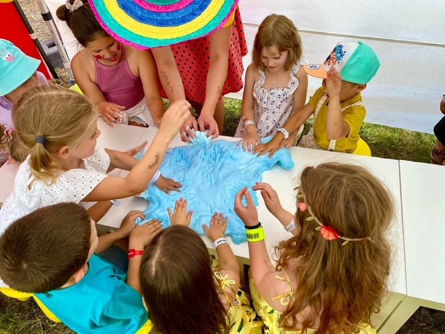 Grupa dzieci zgromadzona wokół stołu, dotykająca dużej, niebieskiej masy plastycznej podczas zabawy na świeżym powietrzu, widoczny fragment kolorowego kapelusza osoby dorosłej.