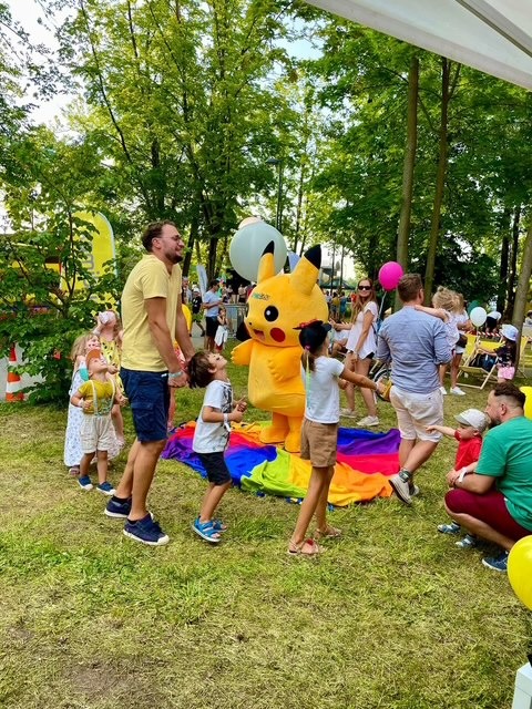 Pikachu w otoczeniu dzieci i dorosłych na trawie, kolorowa chusta animacyjna, plenerowa impreza z animatorem.