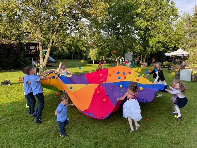 Grupa dzieci bawi się kolorową chustą animacyjną, podrzucając w górę kolorowe piłeczki na trawniku podczas imprezy plenerowej.