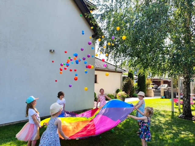 Grupa dzieci bawi się kolorową chustą animacyjną, podrzucając w górę kolorowe piłeczki na tle trawnika i białego budynku.