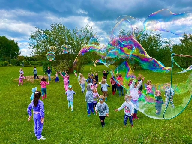 Grupa dzieci bawi się na trawie, otoczona ogromnymi, tęczowymi bańkami mydlanymi tworzonymi za pomocą specjalnych kijów, w tle drzewa i pochmurne niebo.