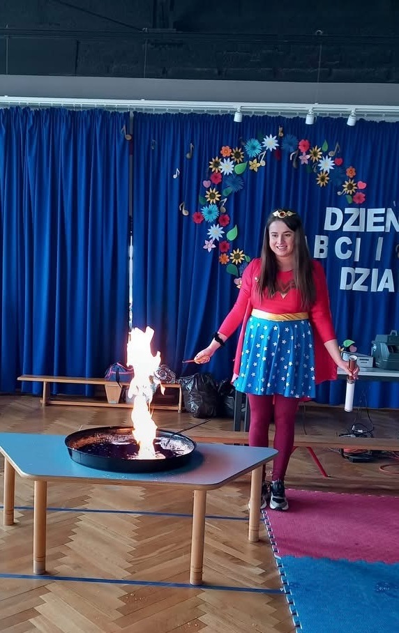 Kobieta w kostiumie Wonder Woman prezentuje pokaz z ogniem w czarnym naczyniu na stole podczas wydarzenia dla dzieci, na tle niebieskich zasłon z dekoracjami kwiatowymi i nutami.