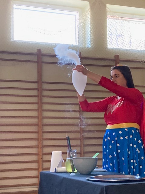 Kobieta przebrana za superbohaterkę w czerwono-niebieskim stroju z gwiazdami, trzymająca balon wypełniony dymem, na stole laboratoryjne naczynia z płynami i akcesoriami, w tle drabinki gimnastyczne.