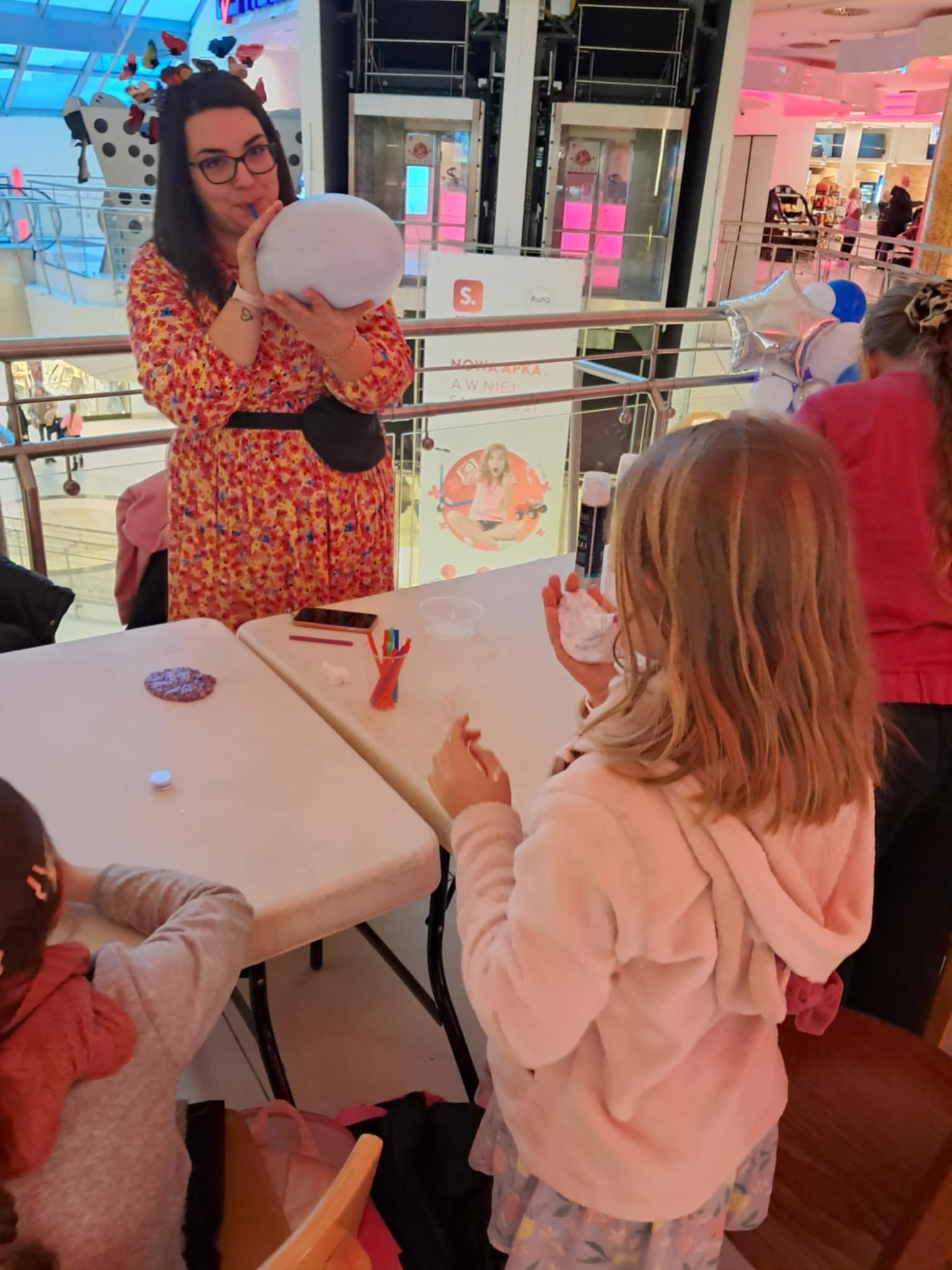 Animatorka w kwiecistej sukience dmucha balon, dzieci przy stolikach bawią się masą plastyczną w centrum handlowym.