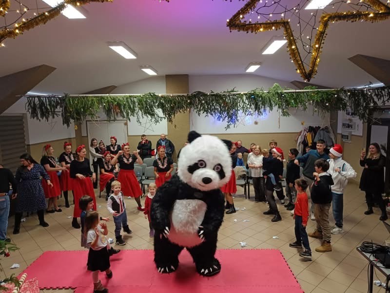 Panda w kostiumie na różowej macie, tancerki w czerwonych spódnicach i dzieci w tle, dekoracje świąteczne na suficie.