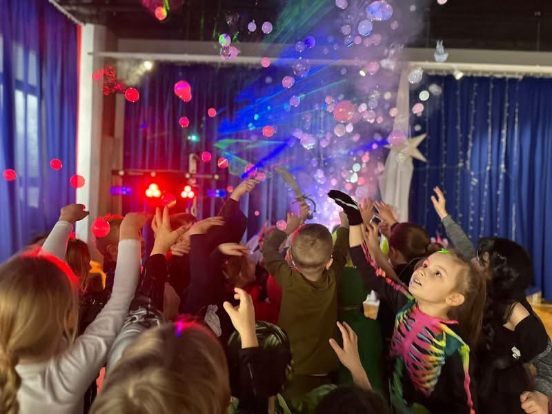 Dzieci z podniesionymi rękami bawią się w dymie i bańkach mydlanych podczas imprezy, oświetlenie sceniczne z laserami i czerwonymi reflektorami, niebieskie zasłony w tle.