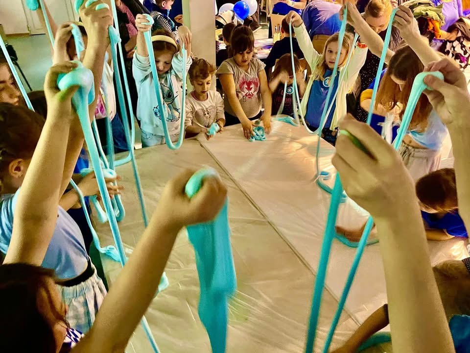 Grupa dzieci bawi się turkusowym slime nad stołem zabezpieczonym folią, ręce uniesione w górę, slime rozciągnięty w długie nitki.