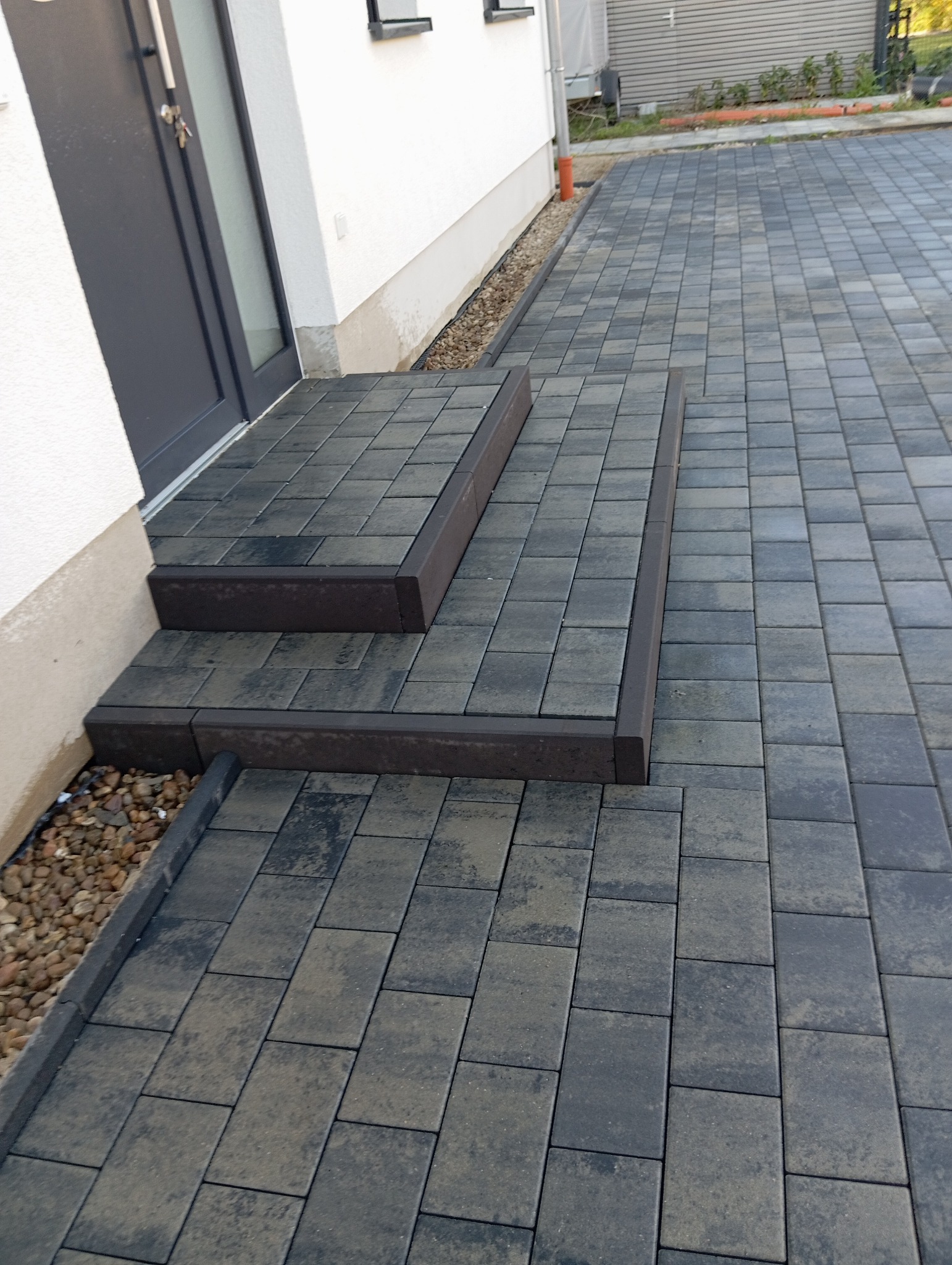 Ułożona kostka brukowa w kolorze grafitowym tworząca schody i chodnik przed budynkiem. Widoczne kamienie ozdobne wzdłuż ściany. Estetyczne wykończenie.