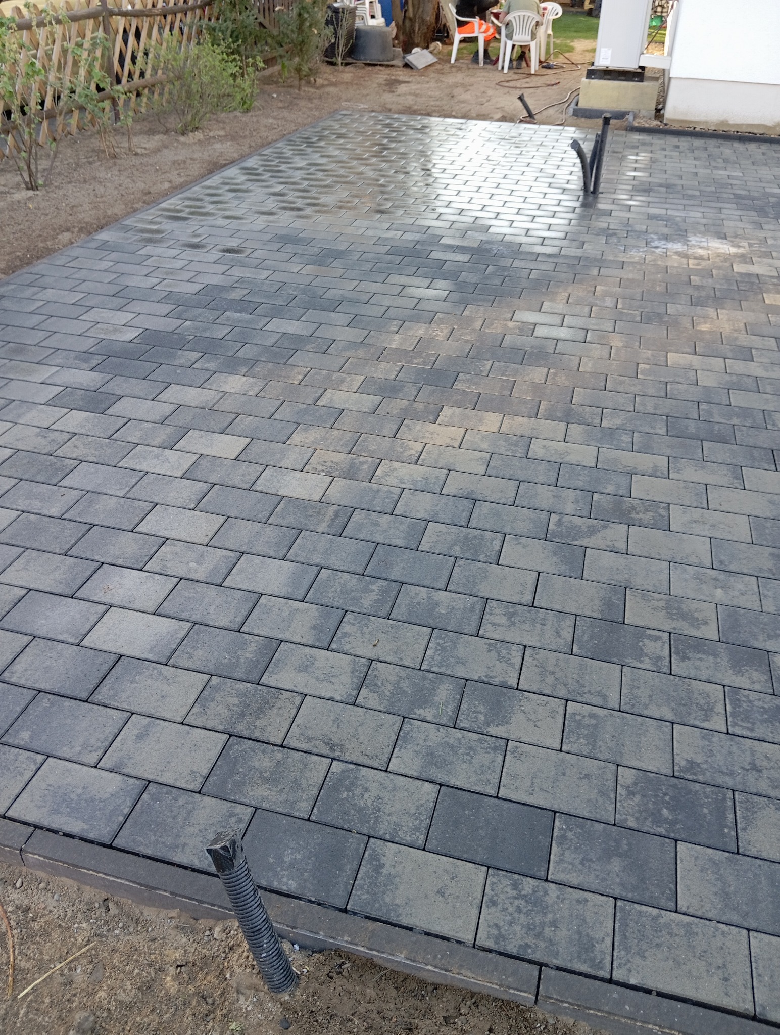 Ułożona, szara kostka brukowa na posesji, widok z góry. Fragment nawierzchni mokry, w tle ogrodzenie z drewnianych sztachet i elementy wyposażenia ogrodu.