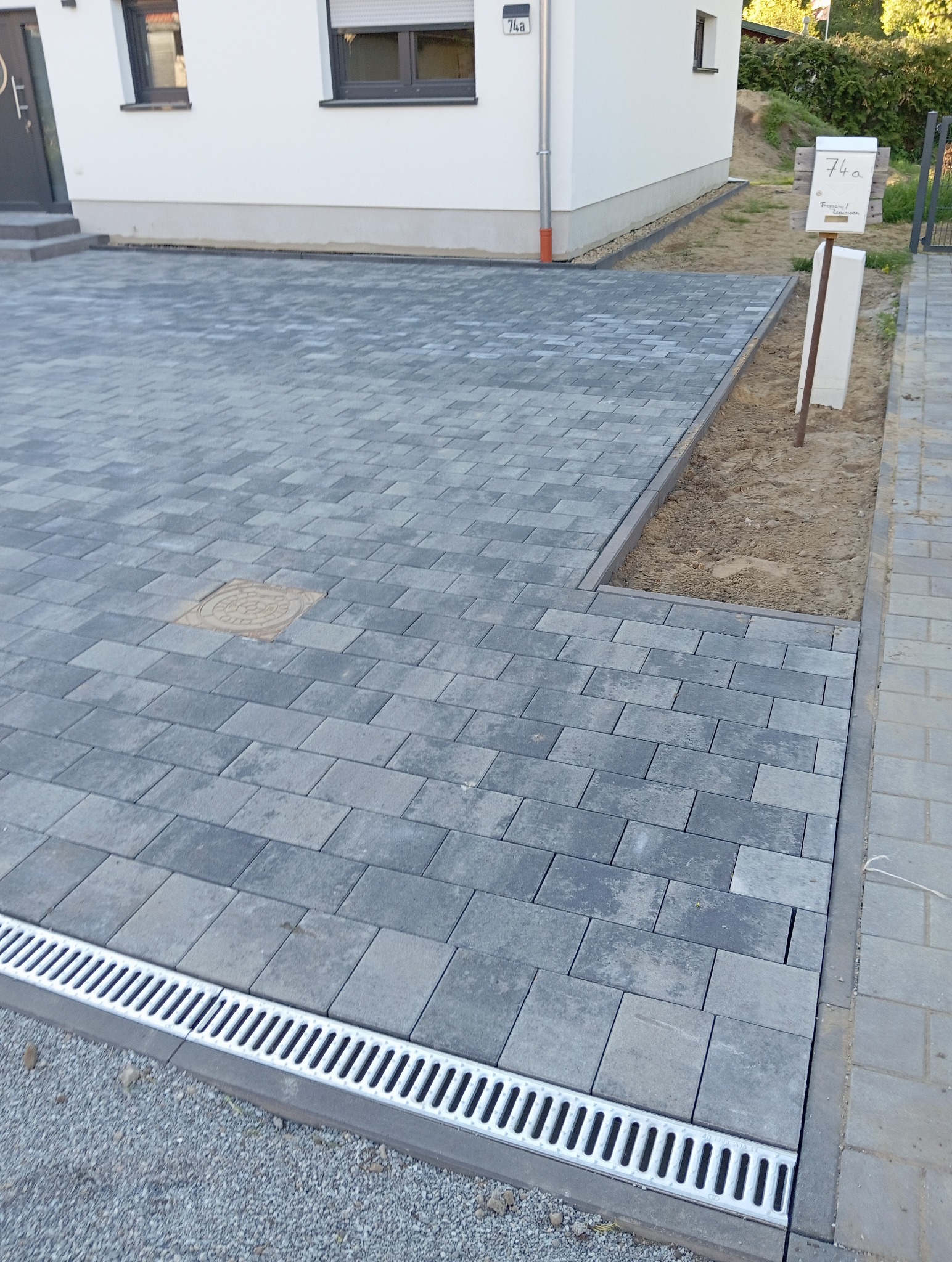 Ułożona, szara kostka brukowa na podjeździe domu z odpływem liniowym i wykończonym obrzeżem. Widoczna skrzynka pocztowa i fragment elewacji budynku.