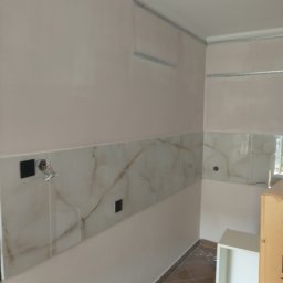 Jakub Łukasik REMONT NA MIARĘ - Wnętrze kuchni w trakcie prac wykończeniowych: położone płytki imitujące marmur na ścianie, widoczne puszki elektryczne i przewody, zamontowane profile do sufitu podwieszanego, częściowo widoczna...