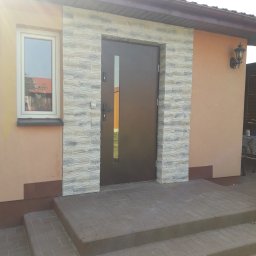 Jakub Łukasik REMONT NA MIARĘ - Wejście do domu z brązowymi drzwiami z wąskim przeszkleniem, obłożone dekoracyjną szarą cegłą, widoczne schody wejściowe z brązowej kostki brukowej, okno po lewej stronie.