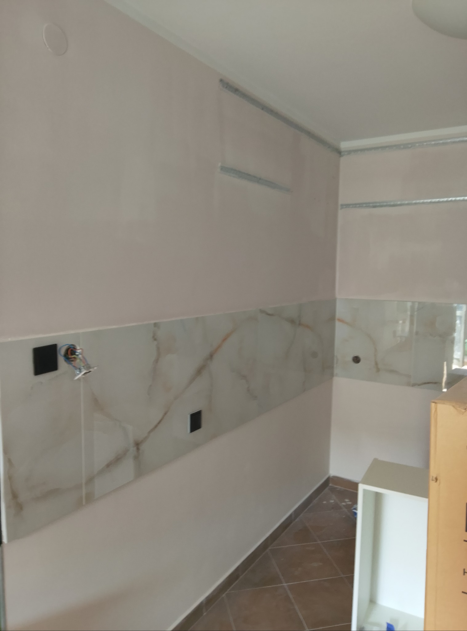 Wnętrze kuchni w trakcie prac wykończeniowych: położone płytki imitujące marmur na ścianie, widoczne puszki elektryczne i przewody, zamontowane profile do sufitu podwieszanego, częściowo widoczna...