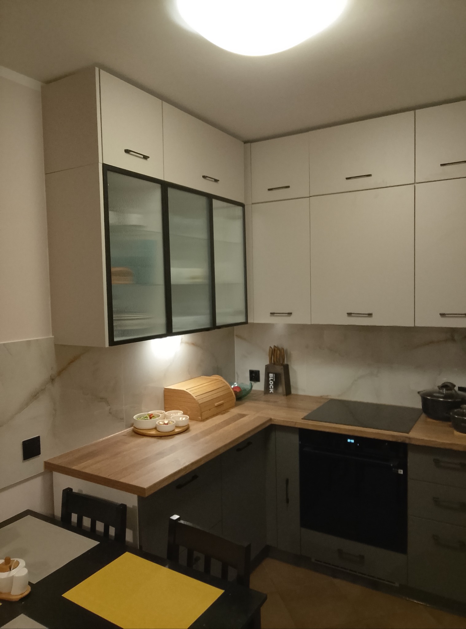 Nowoczesna kuchnia z białymi szafkami górnymi z czarnymi uchwytami, szklanymi frontami i drewnianym blatem, na którym stoi chlebak i miseczki. Na blacie kuchennym płyta indukcyjna i naczynia.