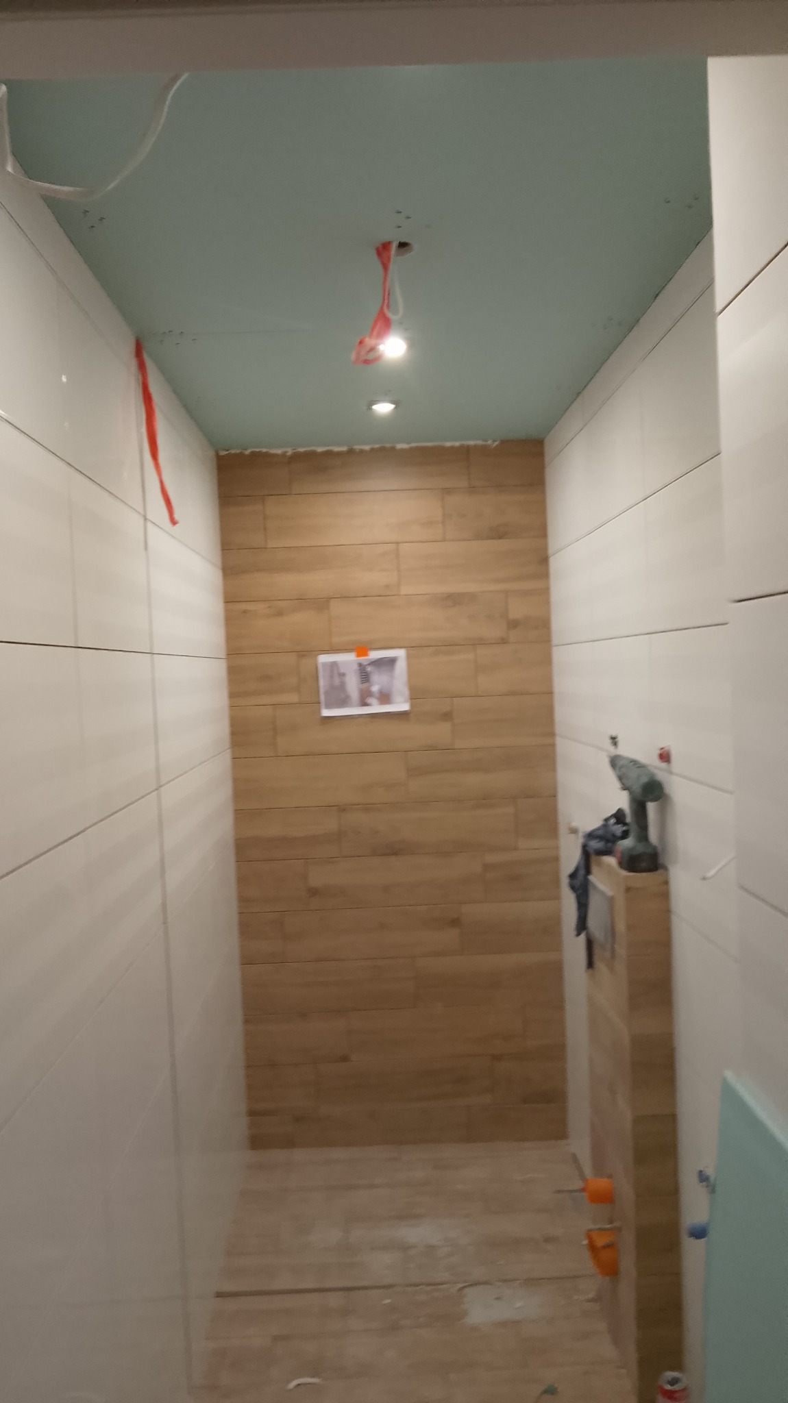 Wnętrze łazienki w trakcie wykańczania: białe płytki na ścianach kontrastują z drewnopodobną okładziną, sufit z płyt kartonowo-gipsowych z widocznymi przewodami i punktowymi lampami, na ścianie...