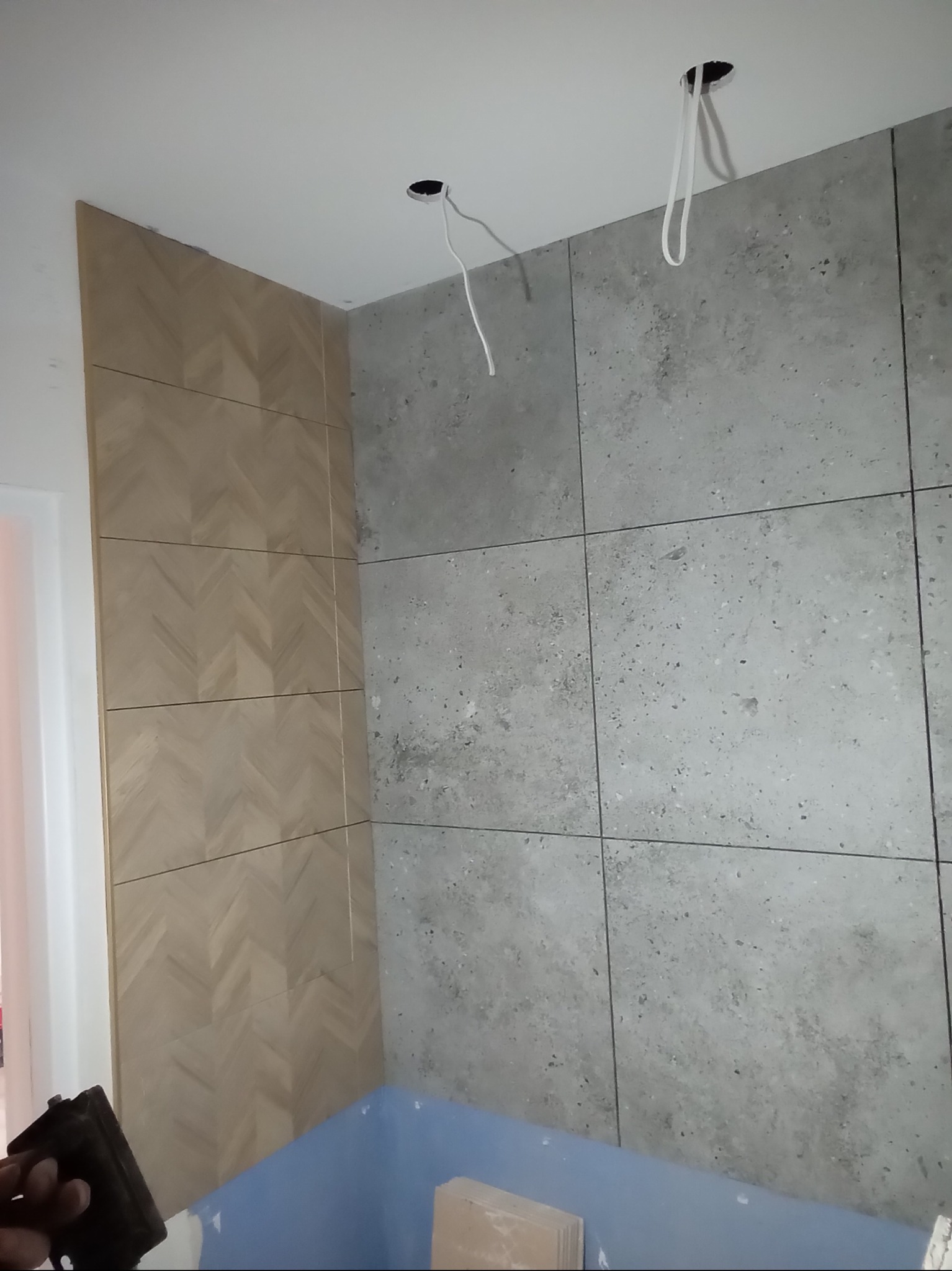 Ściana łazienki wykończona płytkami w dwóch stylach: szare, kwadratowe płytki imitujące beton oraz drewnopodobne płytki ułożone w jodełkę, widoczne przewody elektryczne wystające z sufitu.