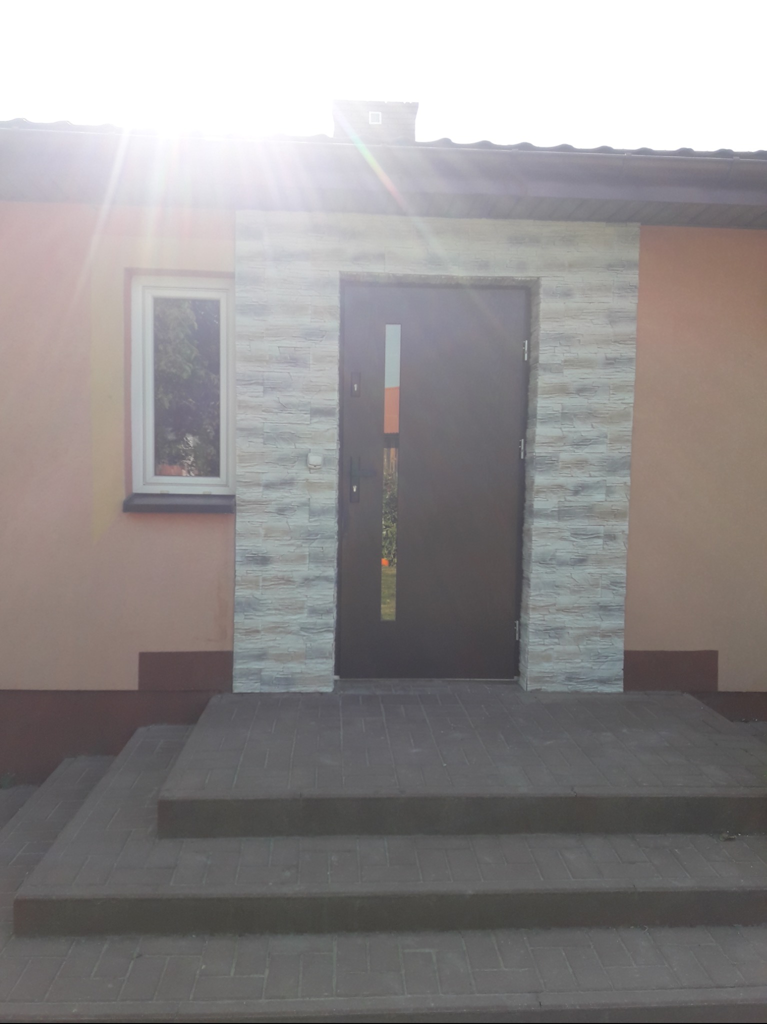Brązowe drzwi wejściowe z wąskim przeszkleniem, otoczone dekoracyjną okładziną z kamienia imitującego cegłę, zamontowane w elewacji domu o jasnoróżowym odcieniu, z widocznymi schodami wejściowymi...