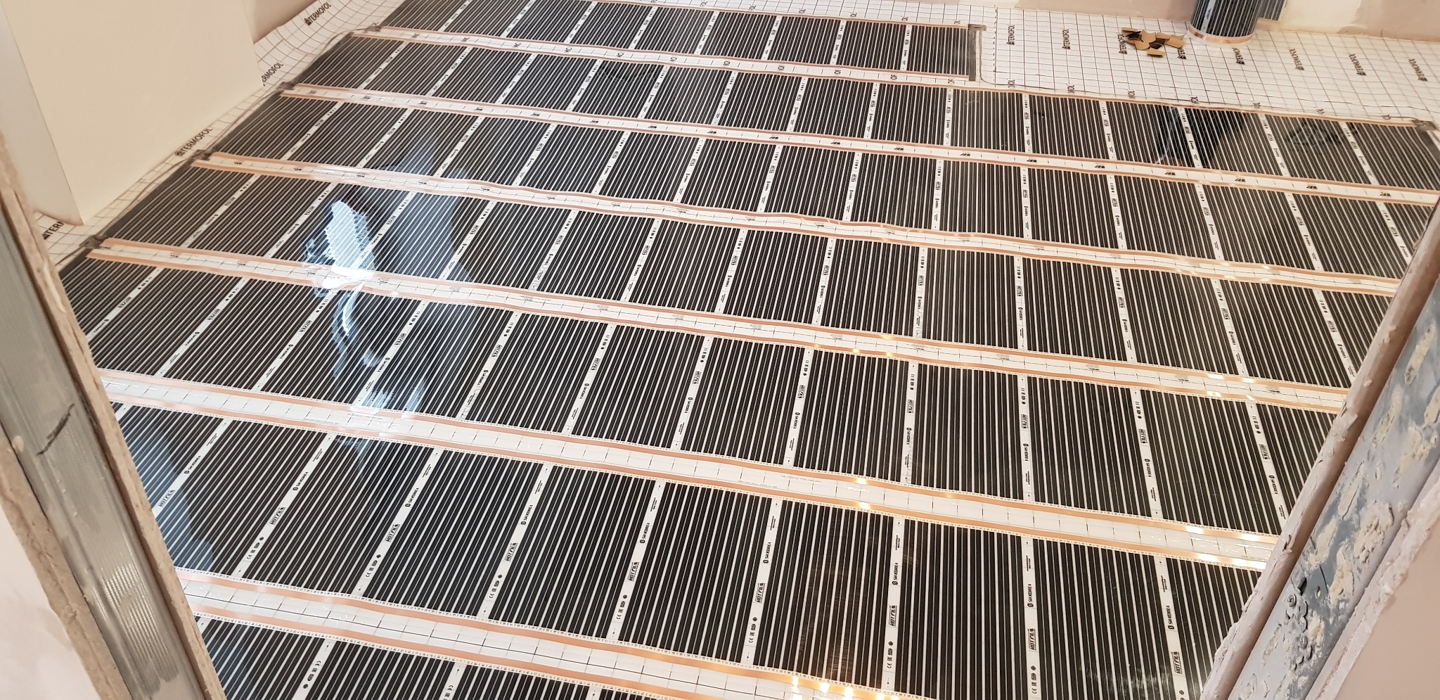 Rozłożone maty grzewcze na podłodze z widoczną siatką i folią termoizolacyjną, przygotowane do dalszych prac wykończeniowych. Widok z góry, ukośnie.