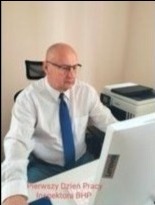 Inspektor BHP przy biurku, w jasnej koszuli i niebieskim krawacie, pracuje na komputerze. Na monitorze widoczny napis 'Pierwszy Dzień Pracy Inspektora BHP'.