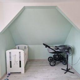 Piotrek Jonienc - Pomalowane na miętowo poddasze ze skośnym sufitem, z białym łóżeczkiem dziecięcym z żyrafą, czarnym wózkiem i stojącym wentylatorem.