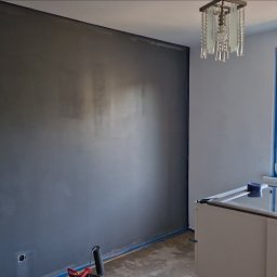 Piotrek Jonienc - Świeżo pomalowana szara ściana w pokoju z zabezpieczonymi taśmą malarską krawędziami, widoczne narzędzia malarskie i otwarte wiadro z farbą, w tle okno z widokiem na blok mieszkalny.