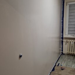 Piotrek Jonienc - Świeżo pomalowana ściana w odcieniach beżu z widocznymi śladami taśmy malarskiej przy listwach i gniazdku elektrycznym, zabezpieczona podłoga z widocznymi śladami farby.