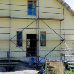 Żółty dom w trakcie ocieplania elewacji, zabezpieczone okna folią, rusztowanie, tablica informacyjna budowy