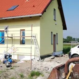 Żółty dom jednorodzinny z czerwoną dachówką w trakcie malowania elewacji, widoczne rusztowanie, okna zabezpieczone folią, teren wokół w trakcie prac wykończeniowych, betoniarka z przodu.