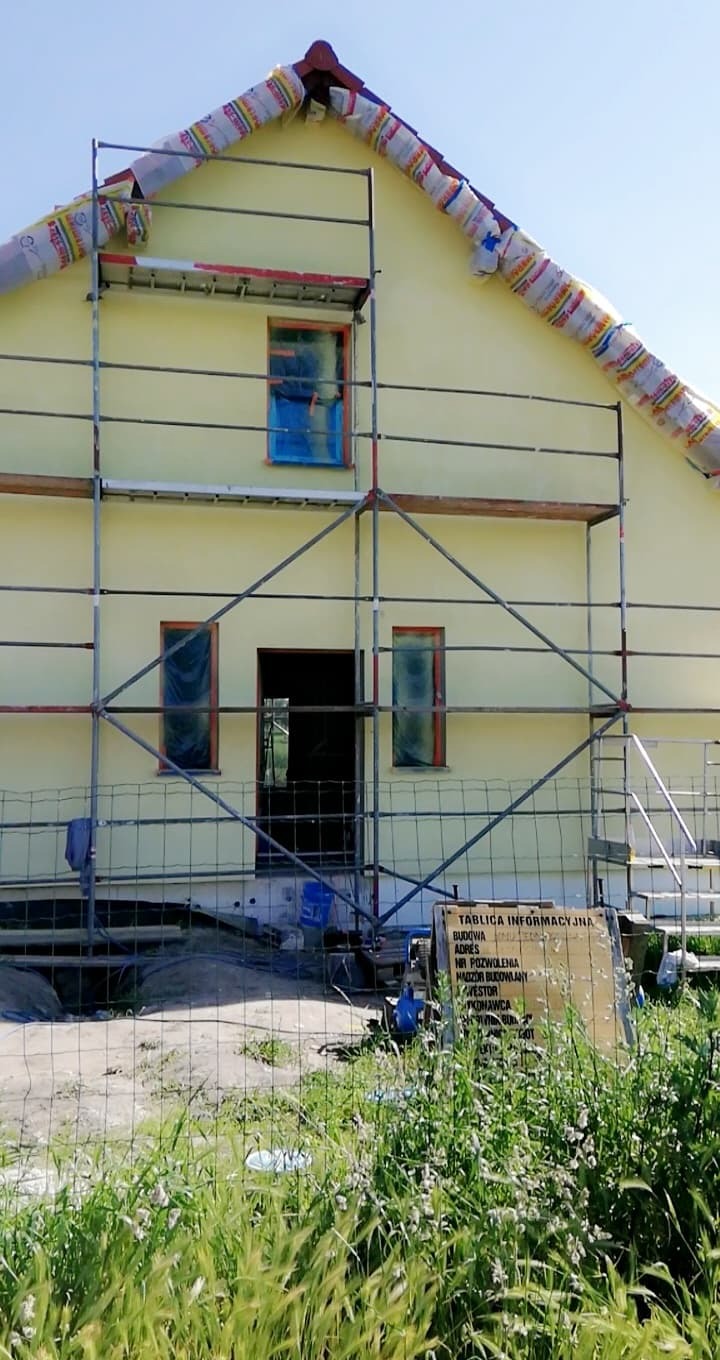 Żółty dom w trakcie ocieplania elewacji, zabezpieczone okna folią, rusztowanie, tablica informacyjna budowy