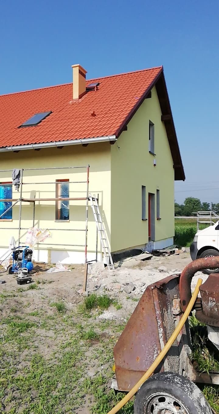 Żółty dom jednorodzinny z czerwoną dachówką w trakcie malowania elewacji, widoczne rusztowanie, okna zabezpieczone folią, teren wokół w trakcie prac wykończeniowych, betoniarka z przodu.