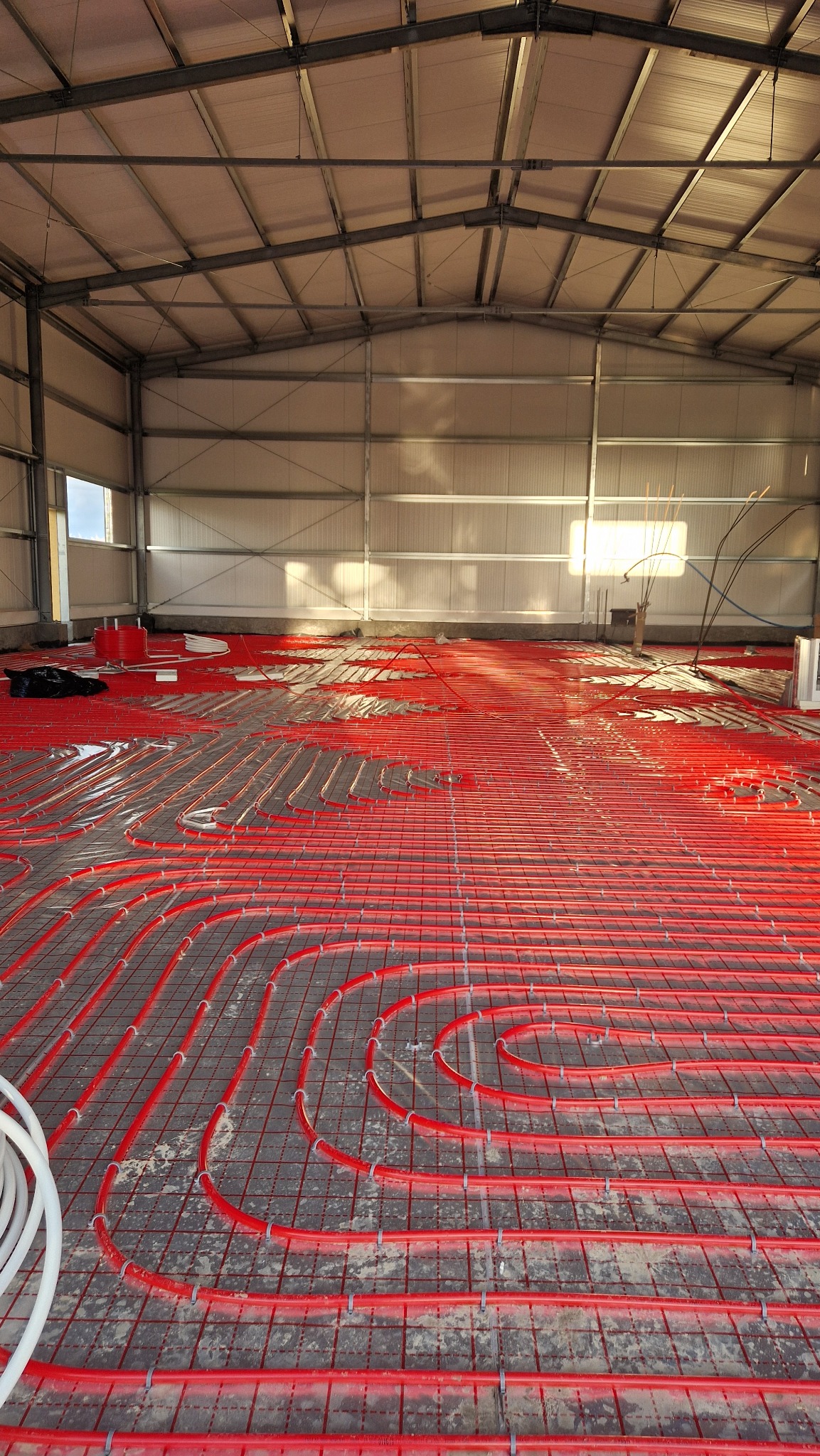 Czerwone rury ogrzewania podłogowego ułożone na siatce w hali przemysłowej, widok z góry, jasne światło wpadające przez okna.