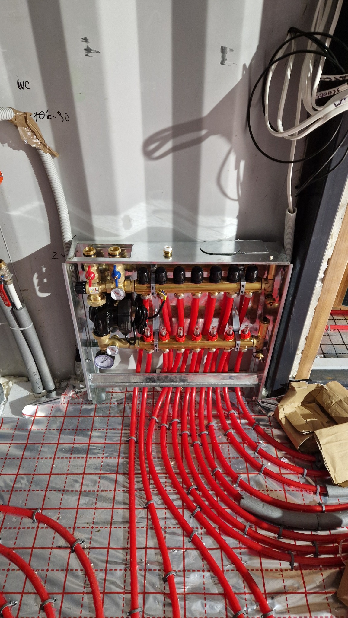 Rozdzielacz ogrzewania podłogowego z czerwonymi rurami na folii izolacyjnej z siatką, widok z góry. Elementy instalacji zamontowane na ścianie.