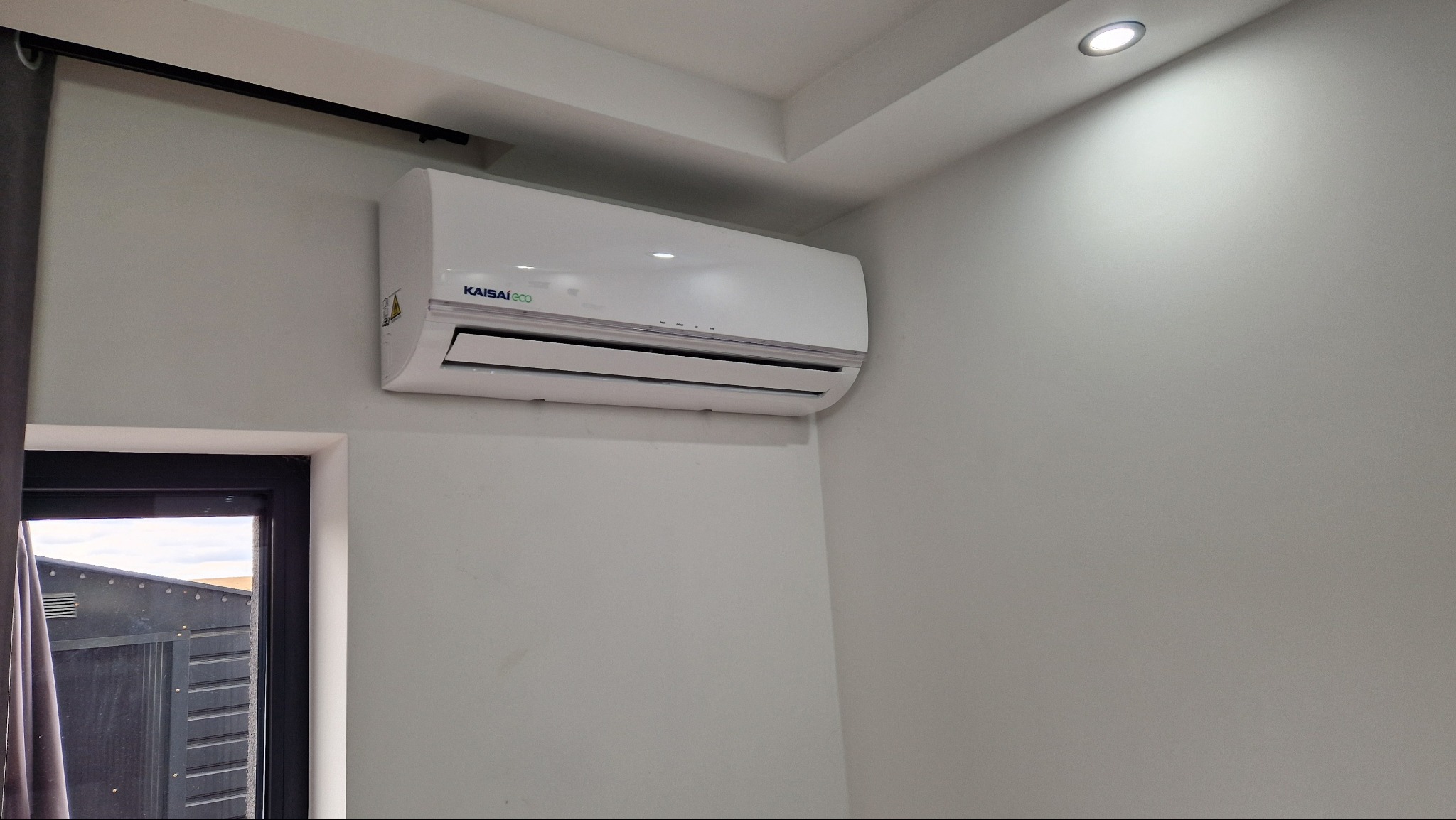 Biała klimatyzacja Kaisaieco zamontowana na ścianie w jasnym pomieszczeniu, widoczna z boku okna z czarną ramą i fragment zasłony. Oświetlenie sufitowe.