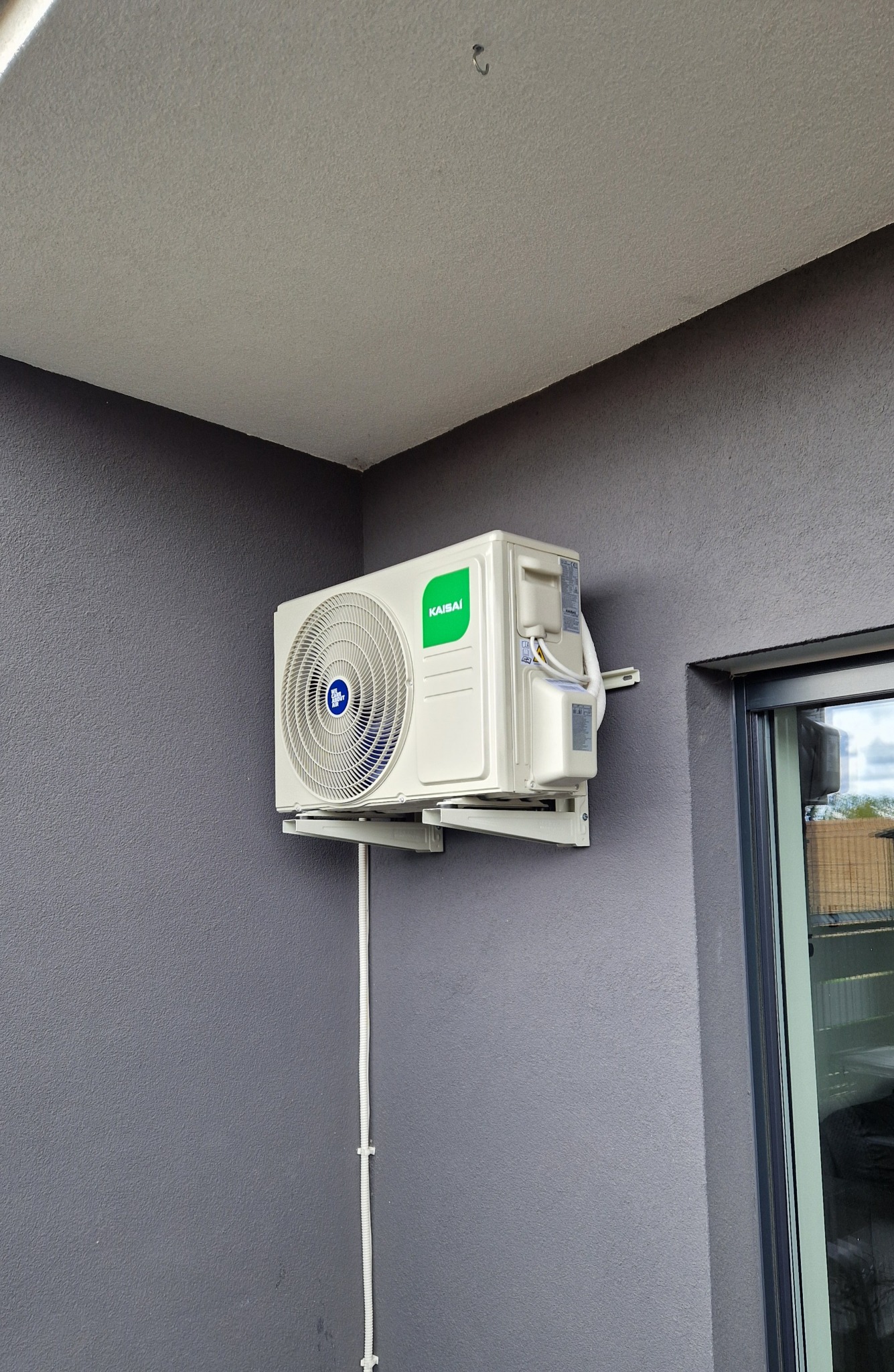Zamontowany klimatyzator Kaisai na szarej ścianie budynku, widoczny kabel w listwie. Obok okno z widocznym odbiciem otoczenia. Ujęcie z dołu, w rogu budynku.