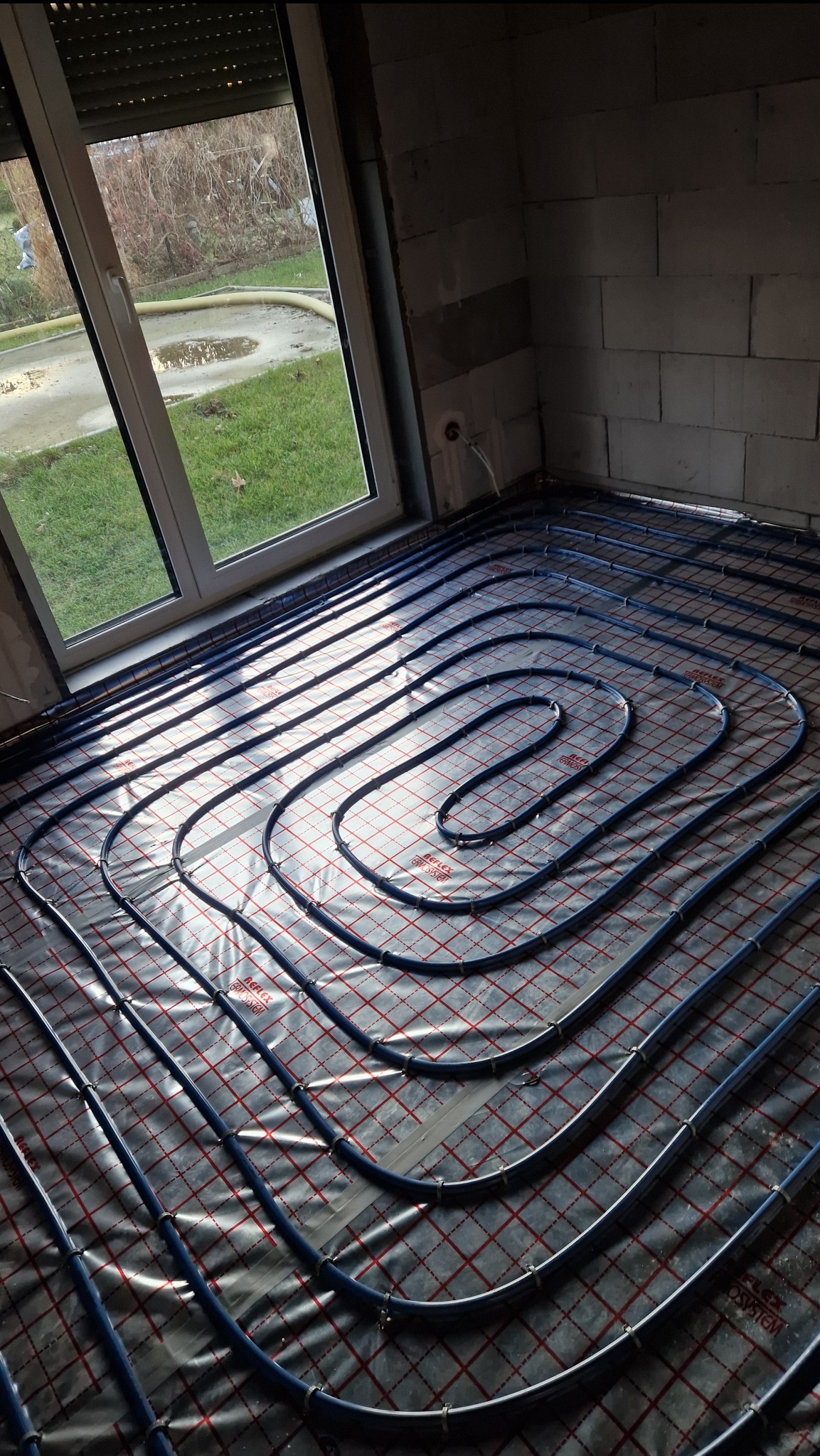 Niebieskie rury ogrzewania podłogowego ułożone spiralnie na folii z czerwoną kratką w surowym wnętrzu z oknem. Widoczne mocowania rur.