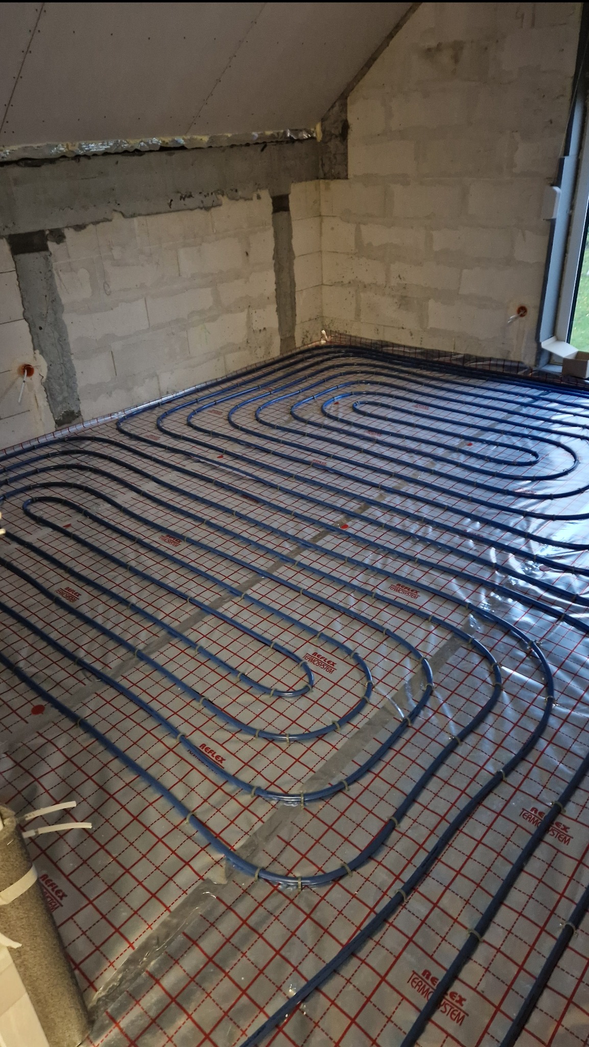 Niebieskie rury ogrzewania podłogowego ułożone na folii z czerwoną kratką w surowym wnętrzu z oknem. Widok z góry, instalacja w trakcie realizacji.