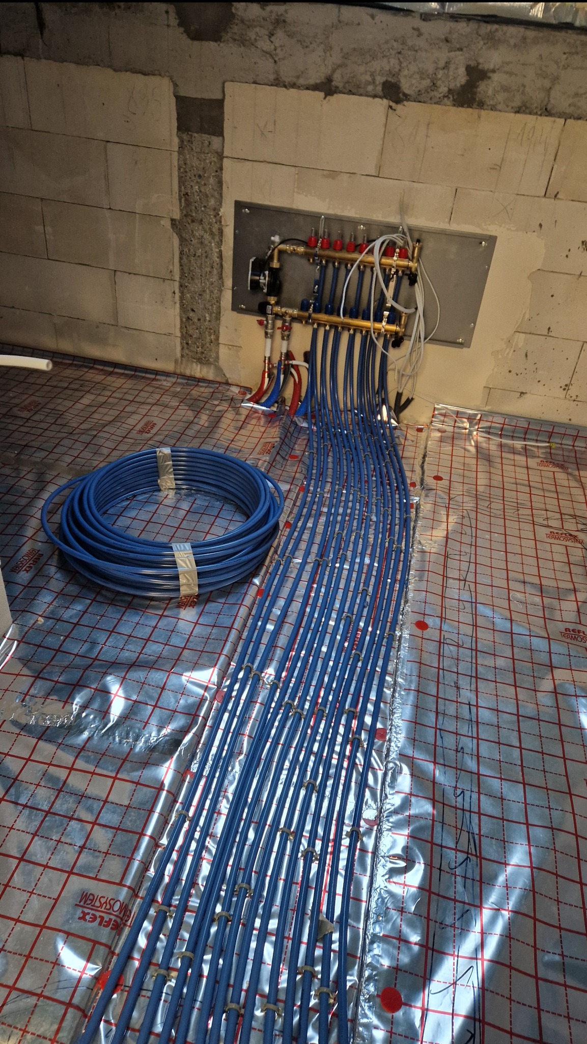 Niebieskie rury ogrzewania podłogowego ułożone na folii refleksyjnej z rozdzielaczem na ścianie z betonu komórkowego. Widoczny zwój rur.