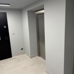 NSB BUILD sp. z o.o. - Jasne, świeżo pomalowane wnętrze z czarnymi drzwiami i wejściem do windy. Minimalistyczny design, gładkie ściany i płytki podłogowe. Oświetlenie sufitowe.